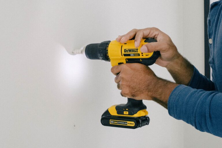 A person uses a yellow and black cordless DeWalt power drill to drill into a white wall, wearing a blue long-sleeve shirt. The drill bit is partially inserted into the wall.