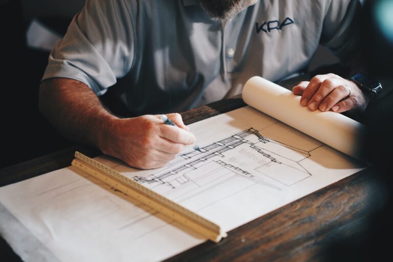 A person in a light gray shirt sketches architectural plans on a large sheet of paper at a desk, using a pencil and a wooden ruler.
