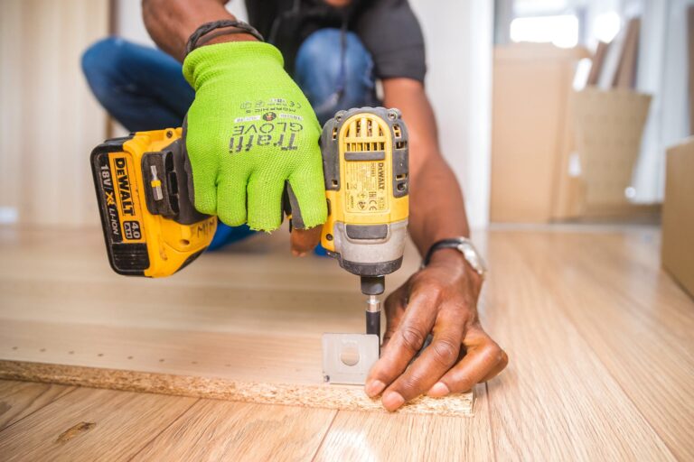 A person wearing a bright green glove uses a yellow cordless drill to assemble a piece of wood furniture on a light wooden floor. Their other hand holds a bracket in place while they work.