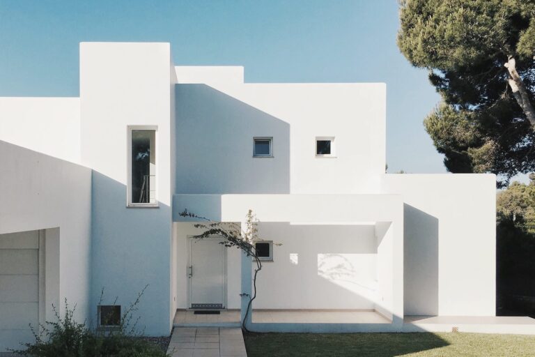 A modern white minimalist house with geometric shapes, flat roof, and large windows, surrounded by a green lawn and trees under a clear blue sky. Shadows create interesting patterns on the building facade.