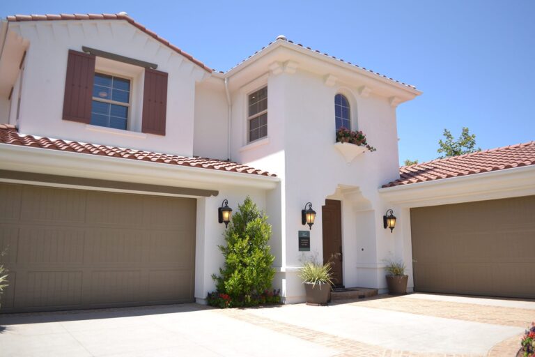 Two-story white house with brown shutters, a red tile roof, and three brown garage doors. There are potted plants and outdoor wall lights near the entrance. The sky is clear and blue.