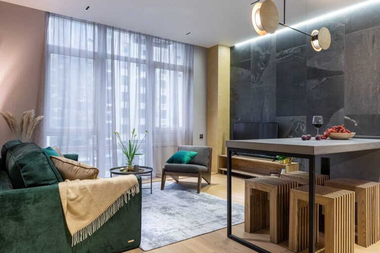 Modern living room with large windows, a green sofa with a beige throw, an armchair, a small coffee table, TV, and a kitchen island with wooden stools. Soft lighting and stylish decor create a cozy atmosphere.