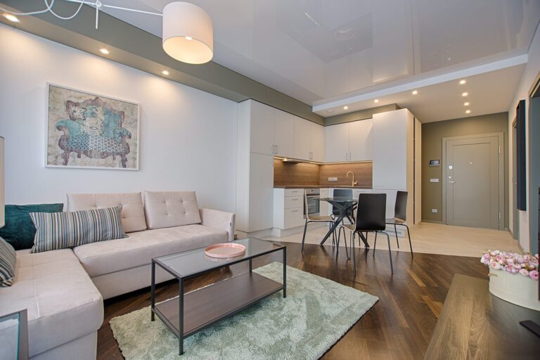 Modern open-plan living room and kitchen with beige sofa, coffee table, dining area with black chairs, light cabinets, wood accents, wall art, and hardwood flooring. Ceiling lights and a rug add warmth to the space.
