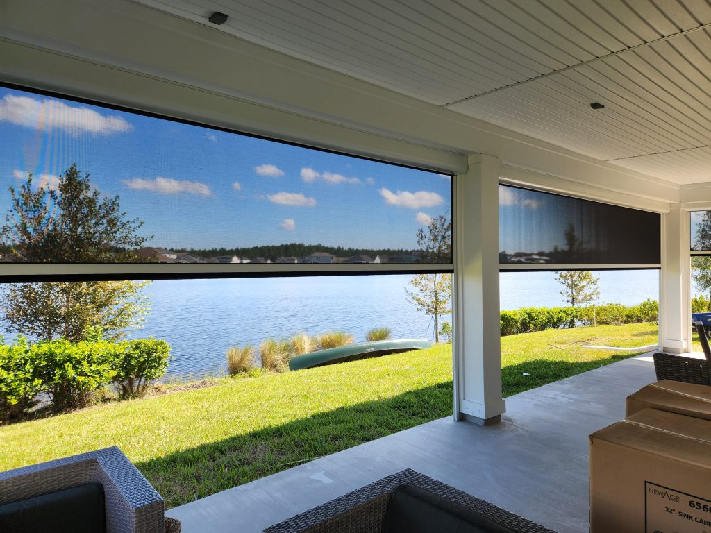 A covered Bradenton FL motorized screen from BGM Wind & Shade overlooks a lake, green grass, bushes, and trees under a blue sky with scattered clouds. Wicker patio furniture and cardboard boxes are visible in the foreground.