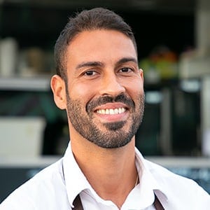 A man with short dark hair and a beard smiles at the camera, wearing a white shirt and standing in what appears to be a modern indoor setting.