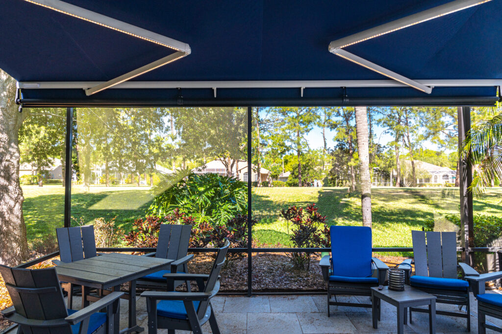 A covered patio features a table with chairs and blue-cushioned seating. The patio is screened, overlooking a green lawn, trees, and landscaped plants on a sunny day.