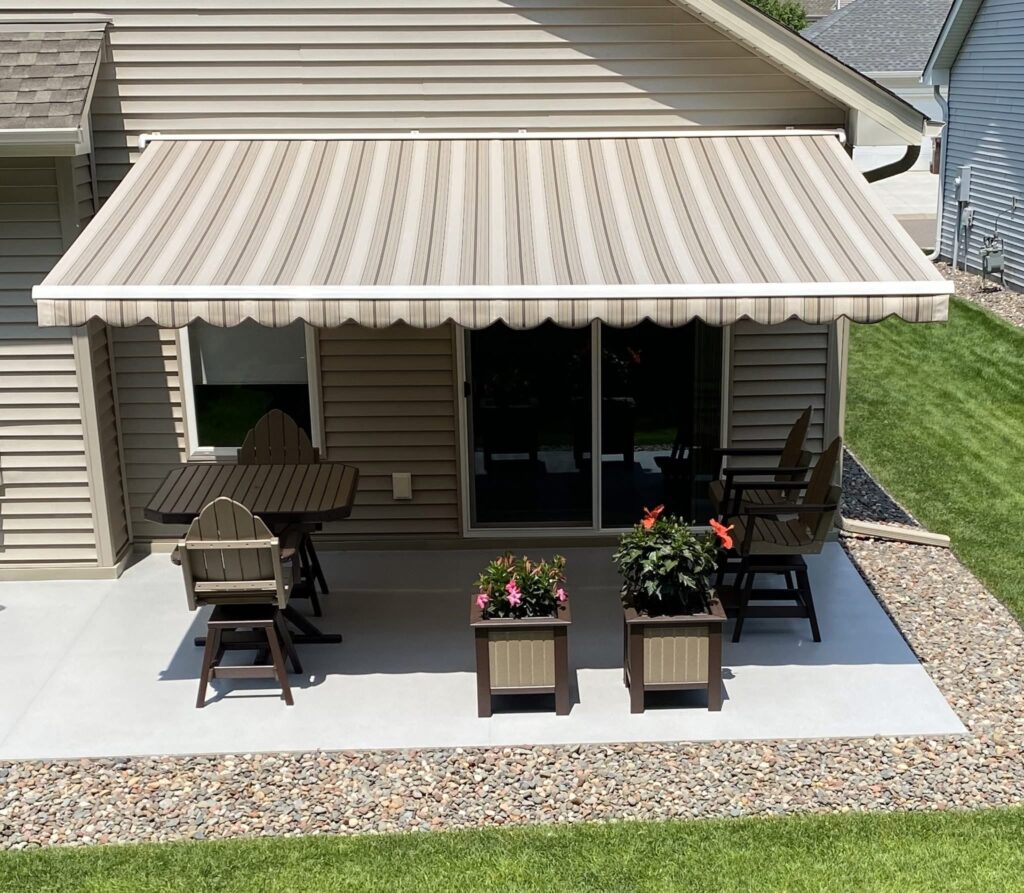 A striped beige retractable awning shades a patio with two planters of flowers, a table with chairs, and a sliding glass door leading into a tan-sided house. The patio is bordered by rocks and surrounded by green lawn.