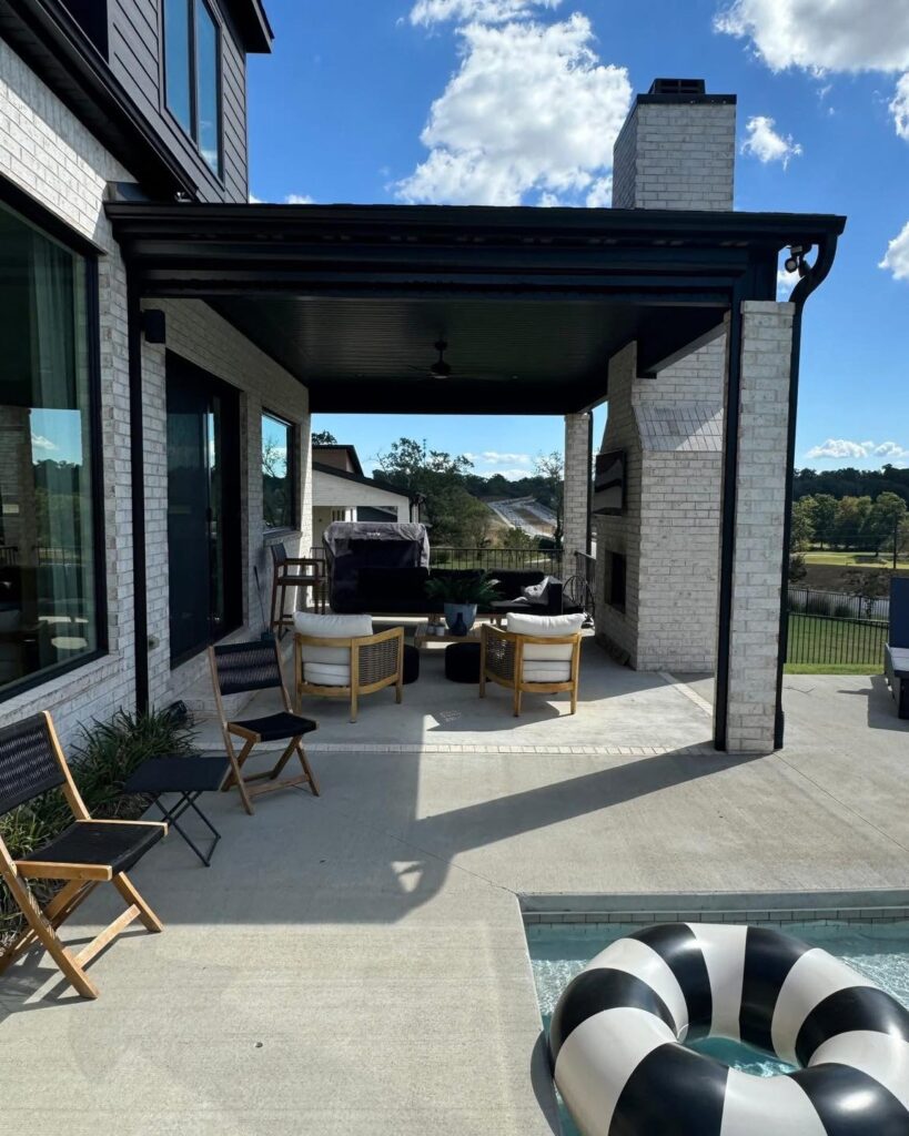 A modern patio with outdoor seating under a covered area beside a pool. There are black and white chairs, a sofa, a small table, and a fireplace. A black and white pool float is in the pool, and trees are visible in the background.