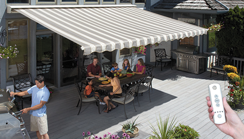A family sits at an outdoor dining table under a striped retractable awning, while a person grills nearby. A hand in the foreground holds a remote control for the awning.