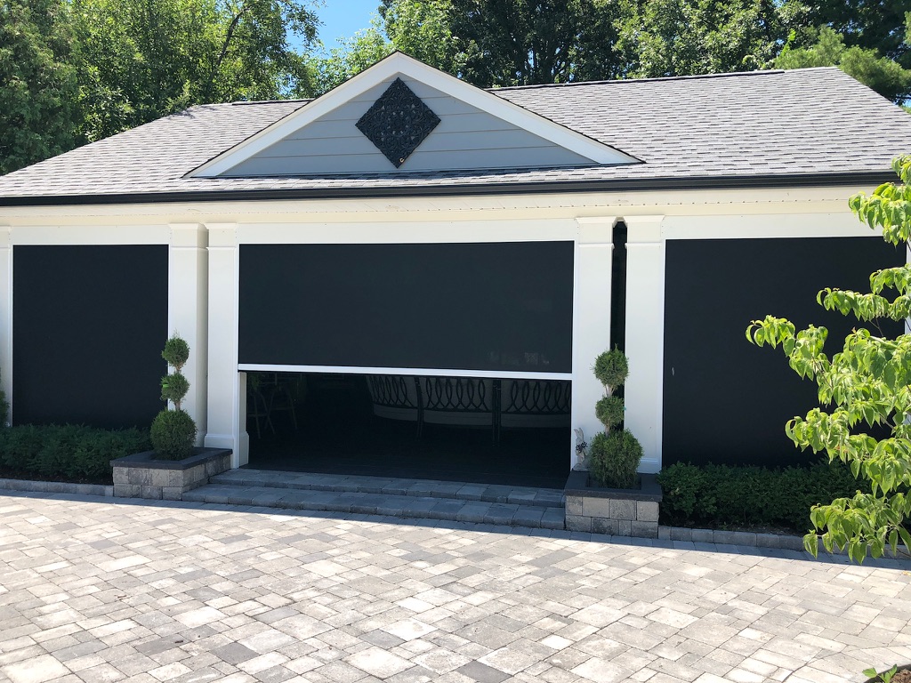A garage with three large black screens covering the entrances, one of which is partially open to reveal a patio swing inside. The building has white pillars, trimmed shrubs, and a paved driveway.