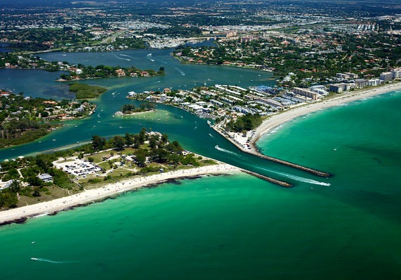 Aerial view of Venice Florida inlet with turquoise water, white sandy beaches, a narrow inlet between two jetties, and boats moving through the channel; urban area and greenery in the background.