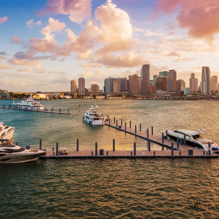 A marina with several yachts docked in the water, set against a city skyline at sunset. Tall buildings are illuminated by warm sunlight, and fluffy clouds fill the colorful sky above.