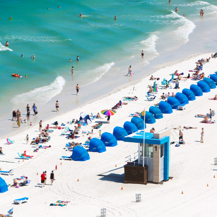 A sunny beach with turquoise water, rows of blue beach cabanas, colorful umbrellas, and many people relaxing on the sand and swimming in the ocean. A white and blue lifeguard station is near the cabanas.