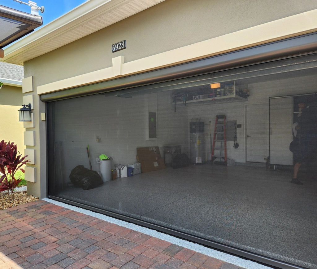 A residential garage with the door open, showing a clean interior with some tools, storage bins, lawn equipment, and a red ladder along the back wall. The driveway is paved with bricks.