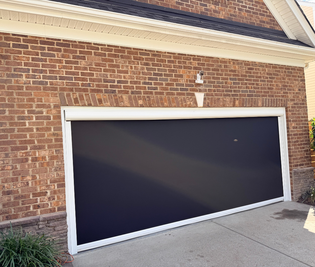 A garage door is partially open, tilted upward with a black covering pulled down behind it. The house exterior is red brick with a white trim and a small light fixture above the garage.