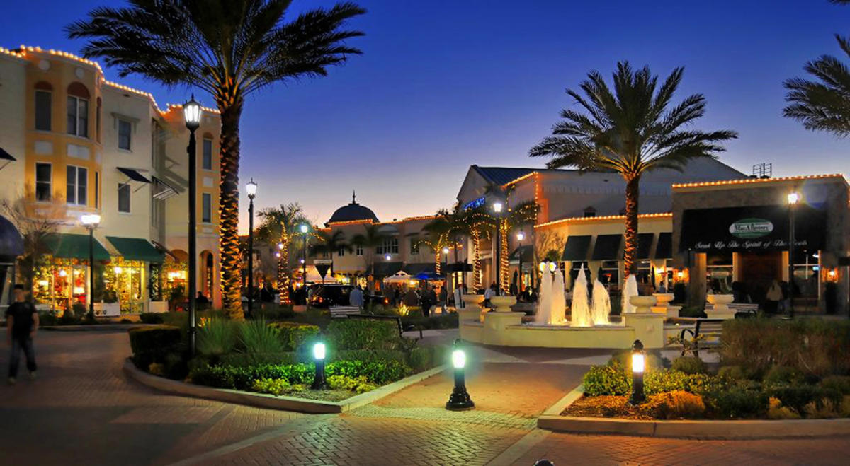 A lively outdoor plaza at dusk with palm trees, glowing lights outlining buildings, a central fountain, and people walking and sitting around, creating a warm and inviting atmosphere.