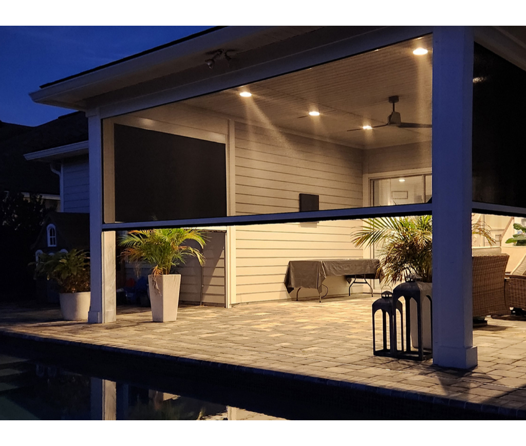 A covered patio at dusk with potted plants, outdoor furniture, ceiling lights, and a view of a pool in the foreground. The area is illuminated by warm lighting, creating a cozy ambiance.